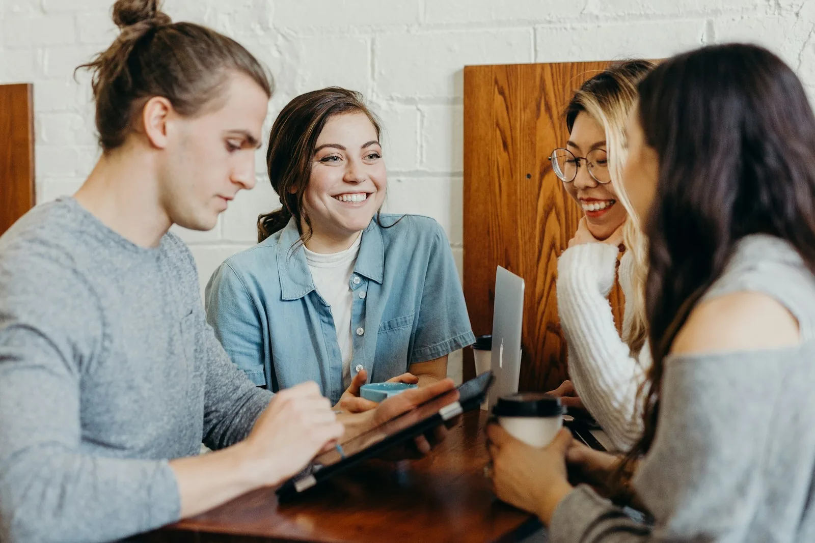 Four young people sitting around a table together with laptops and coffee, talking and laughing